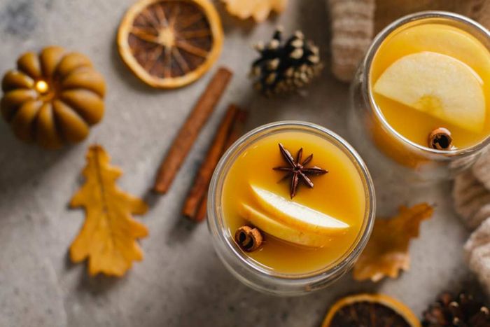 A top-down view of two glasses of apple cider adorned with apple slices and star anise, alongside cinnamon sticks on a table.