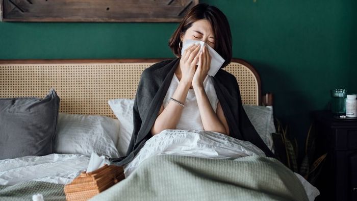 A young Asian woman lying in bed, using a tissue to blow her nose.