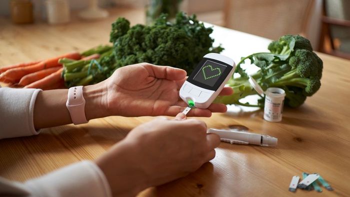 A young woman is seated at a table, using a glucometer to check her blood sugar levels while various vegetables are spread across the surface.