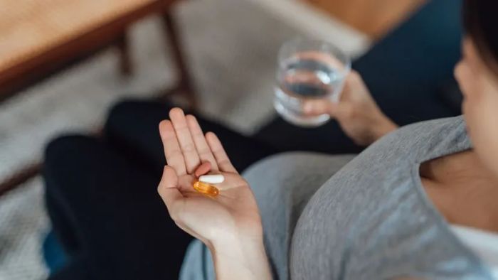 A pregnant woman gently holds her prenatal vitamins in her left hand while sipping from a cup of water in her right hand.