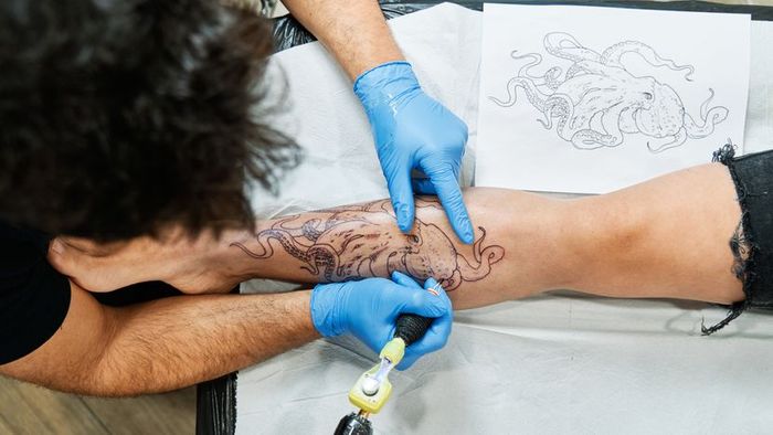 A tattoo artist working on an octopus design on a person’s leg.