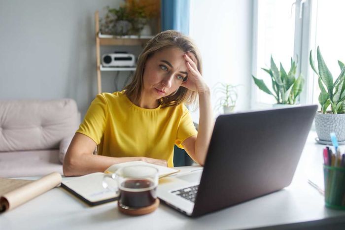 A woman wearing a vibrant yellow jacket sits in front of a laptop, visibly stressed.