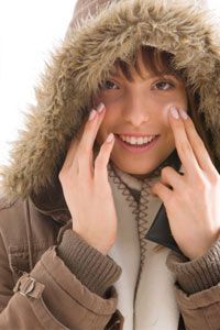 A cheerful young woman in a fur-lined hooded jacket applying moisturizer to her cheek