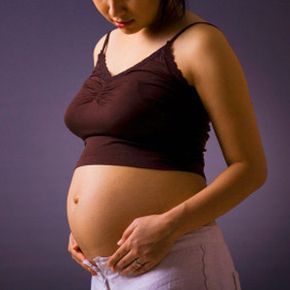 A pregnant woman stands against a grey backdrop, her stomach visibly exposed in a studio setting
