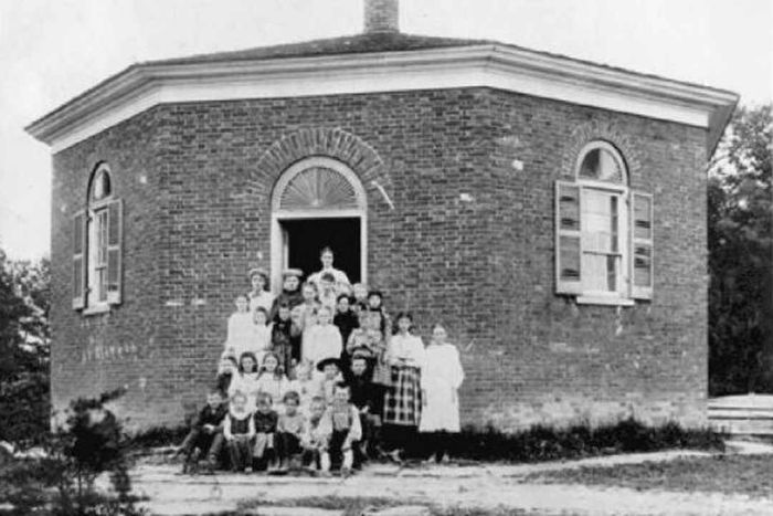A group of students and their teacher standing outside a one-room schoolhouse.