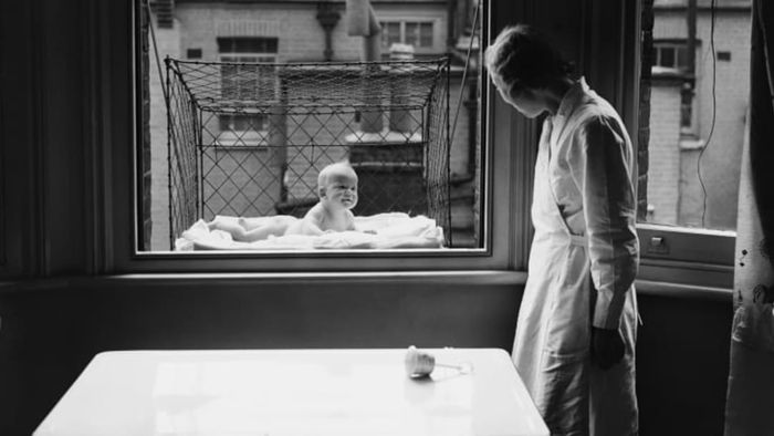 A nanny watching over a baby in a wire cage hanging outside a high-rise tenement window.