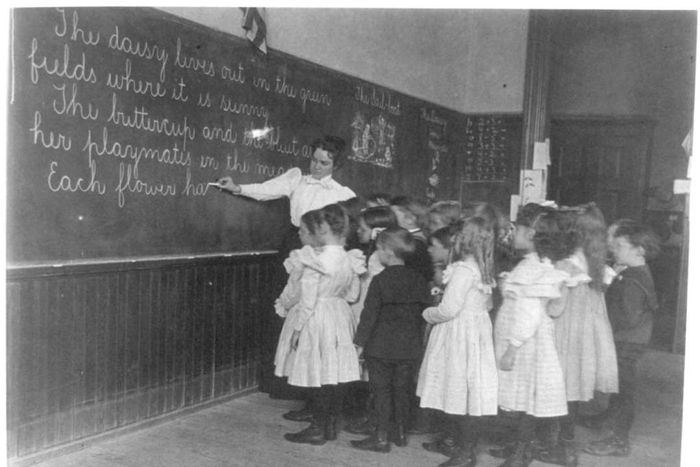 Elementary school students standing and observing their teacher write on a blackboard