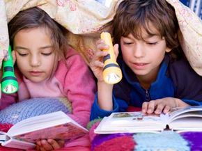A boy and girl reading by flashlight beneath their blankets