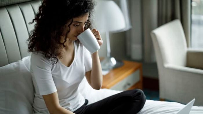 woman sipping from a cup while using a laptop on a hotel bed