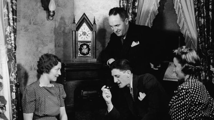 Four men and women gather closely together while listening to their home radio console, 1930s.