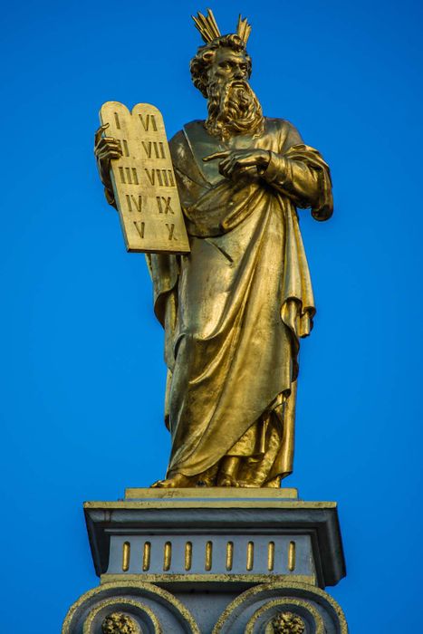 A golden statue of Moses holding the Ten Commandments, with horn-like rays of light radiating from his head.