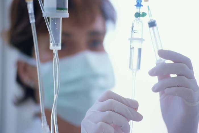 A healthcare professional wearing a mask administers medication through an IV tube.