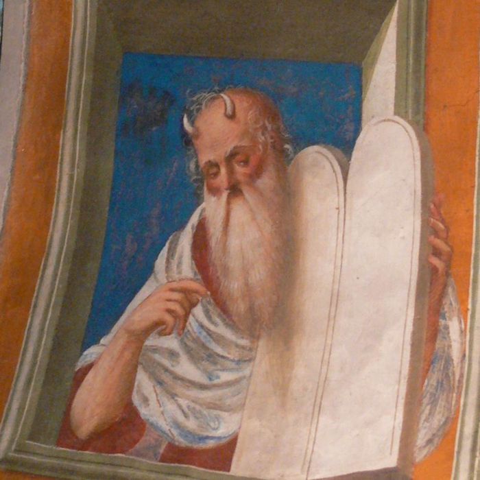 A depiction of Moses, mostly bald with a long white beard and two small horns, gesturing toward the blank tablets he holds.