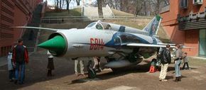 The MiG-21, a white, green, and red Soviet fighter jet, is surrounded by groups of people examining it.