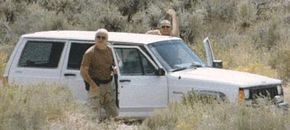 Two men clad in brown shirts and camouflage pants stand beside a white SUV.
