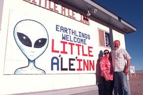 A couple stands before a mural that reads 'Earthlings Welcome' and 'Little A'Le'Inn'.