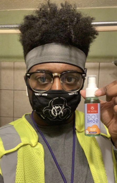 A reviewer image of a person wearing clear glasses with their mask on, holding up the anti-fog spray