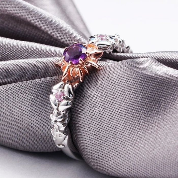 The ring displayed on a gray napkin, showcasing a central amethyst, a sunburst setting, and a band adorned with a braided floral design