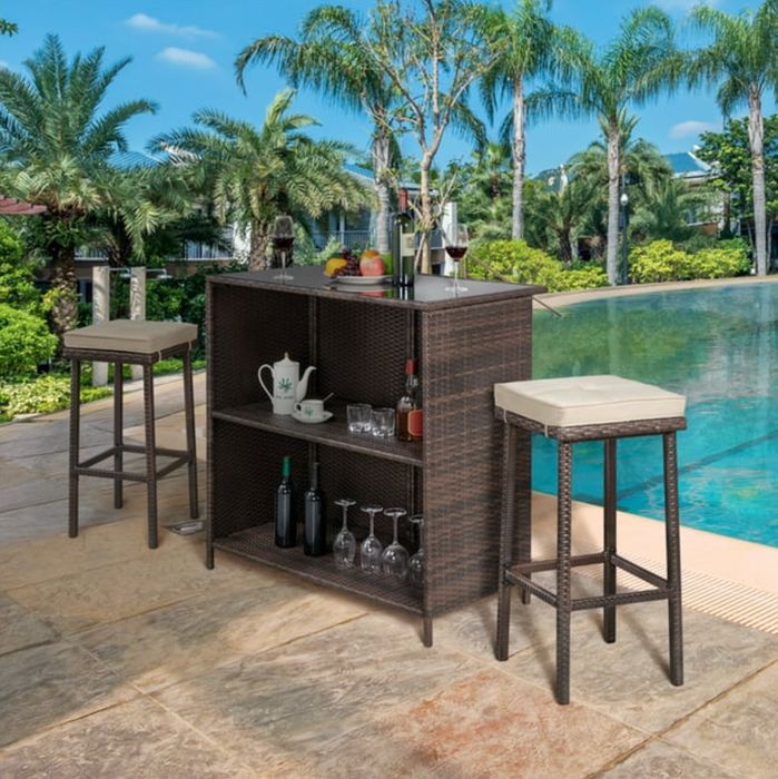 the dark rattan bar table and stools with tan cushions set against the backdrop of a beautifully decorated pool deck