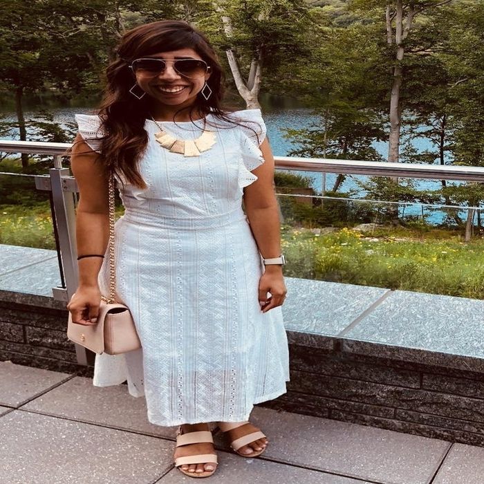 A reviewer wears a sleeveless white dress accessorized with a necklace, sunglasses, and a handbag, standing near a railing with a natural backdrop.