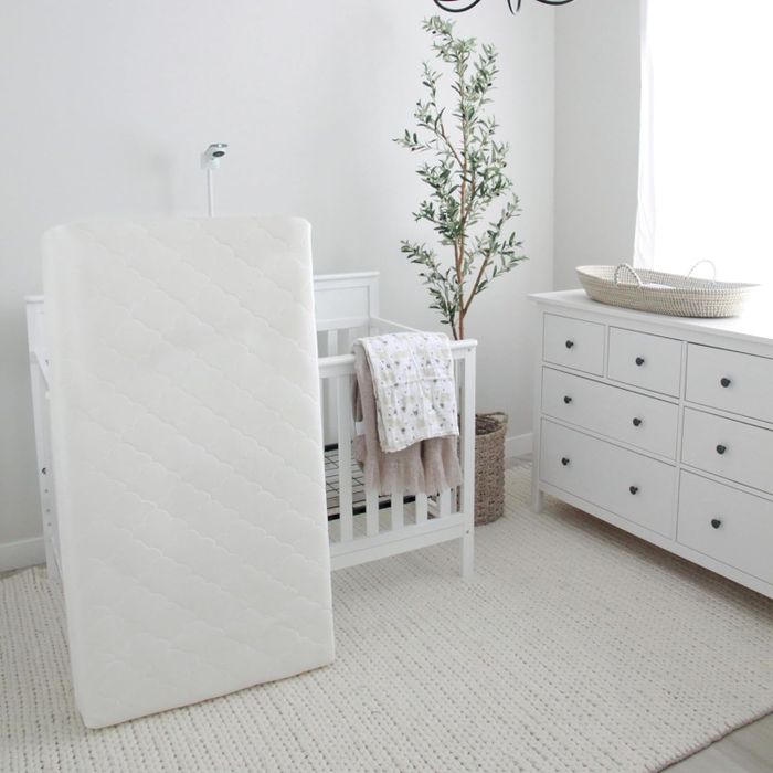 A crib mattress propped against a wall beside a white crib and dresser in a nursery