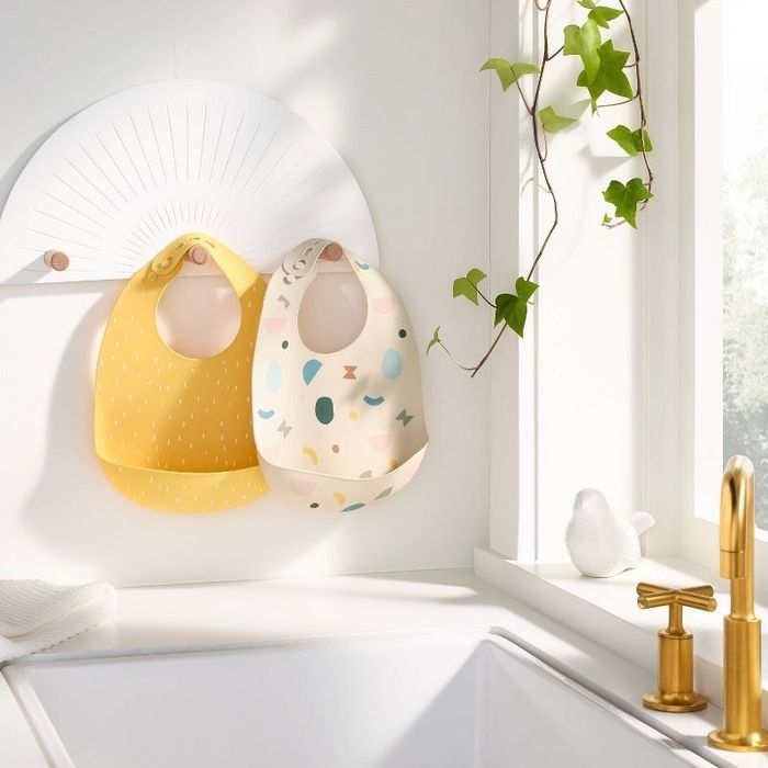 Two bibs, one yellow with white polka dots and the other white with vibrant shapes, hang in a cheerful kitchen next to a gold faucet