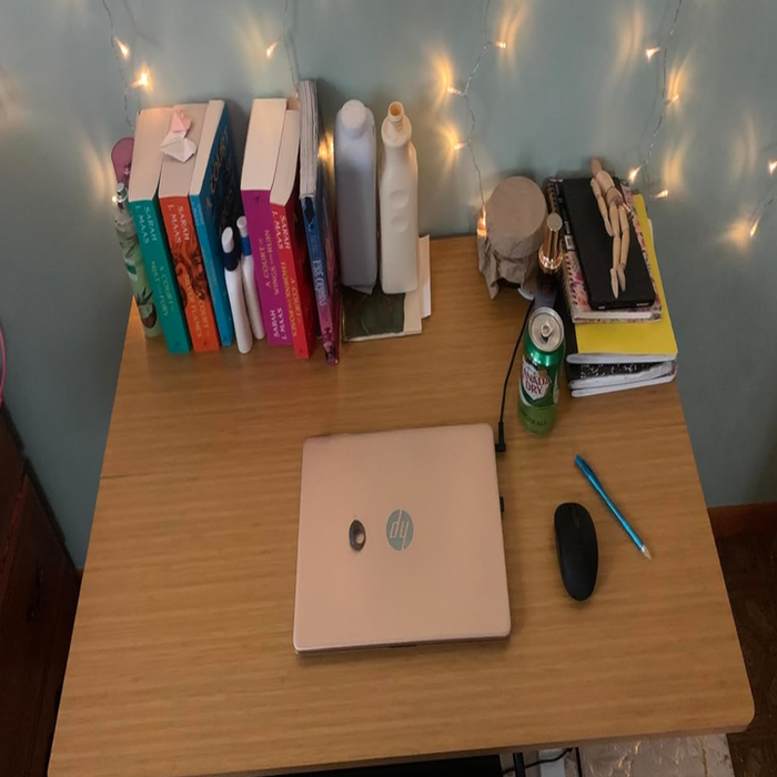 A tidy wooden desk adorned with books, a laptop, a mouse, a soda can, notebooks, a pen, skincare products, and decorative fairy lights
