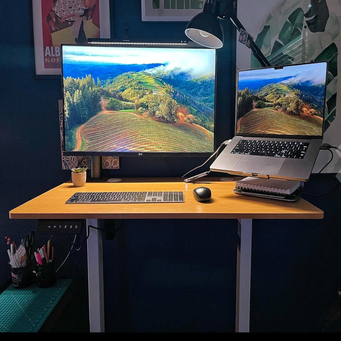 A home office setup with a spacious desk holding a large monitor, a laptop showing a scenic image, a keyboard, mouse, lamp, and various office essentials