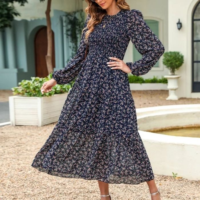 A model in a floral-patterned maxi dress with long sleeves, standing outdoors by a fountain.