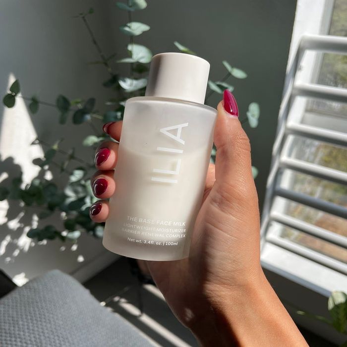 A hand holding a bottle of ILIA The Base Face Milk, with soft sunlight filtering through greenery in the background.