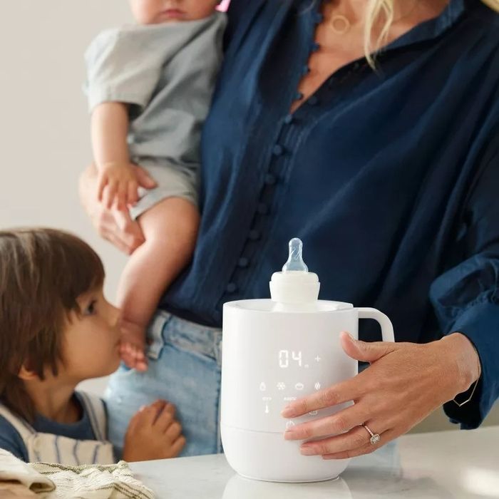 A person holding a baby, with a toddler nearby, uses a sleek white digital bottle warmer. The focus is on the appliance in use