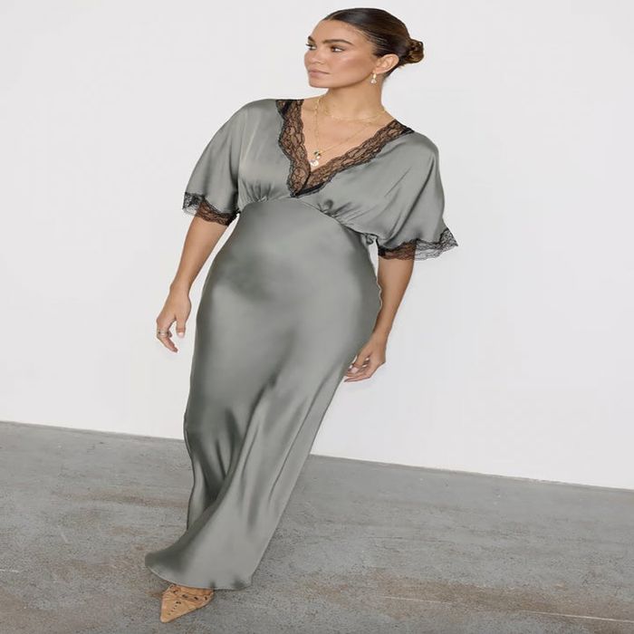 A person wears a refined satin dress featuring lace trim, paired with a gold necklace and a chic bun hairstyle, standing against a neutral backdrop