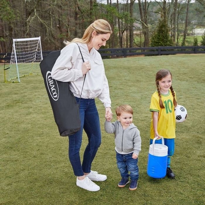 An adult and two children stand at a soccer field, with a Graco bag nearby and a child holding a soccer ball and a cooler. They are dressed casually for an outdoor activity