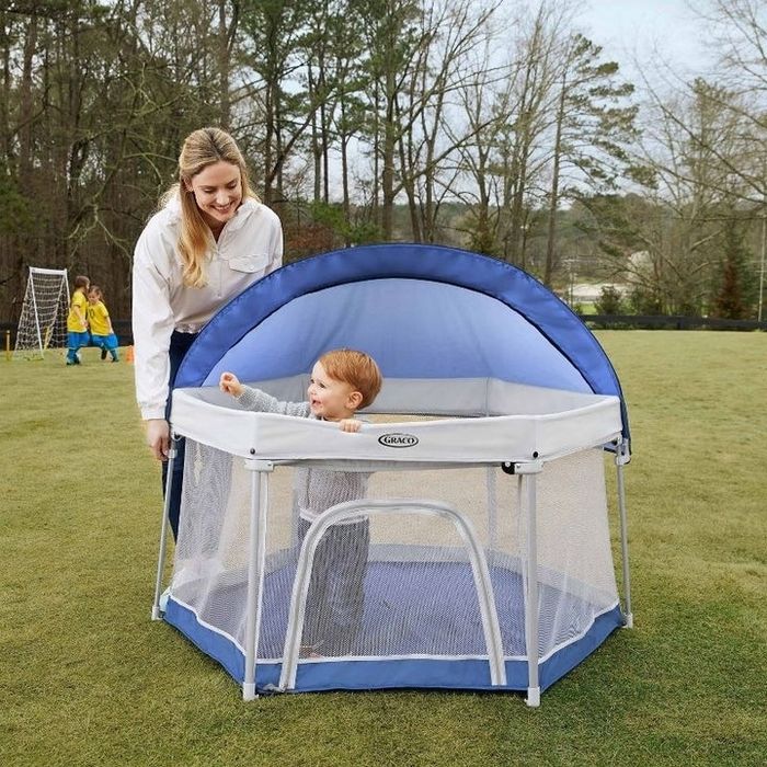 An adult smiles at a baby inside a shaded playpen. The outdoor setting includes trees and a field in the background