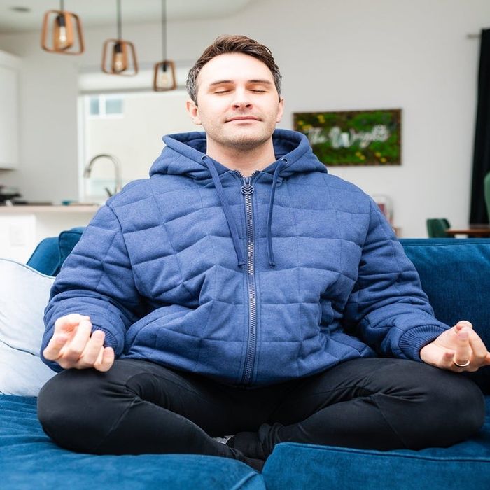 A person meditates on a sofa, wearing a cozy quilted jacket with a hood and black pants in a relaxed home environment