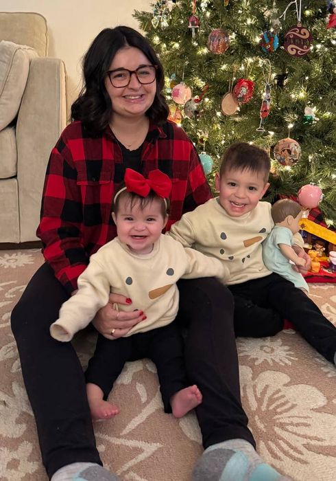 A family gathered around a Christmas tree; the adult is wearing a plaid shirt, the children sport snowman shirts, with one child wearing a hair bow. A cozy, festive holiday moment.
