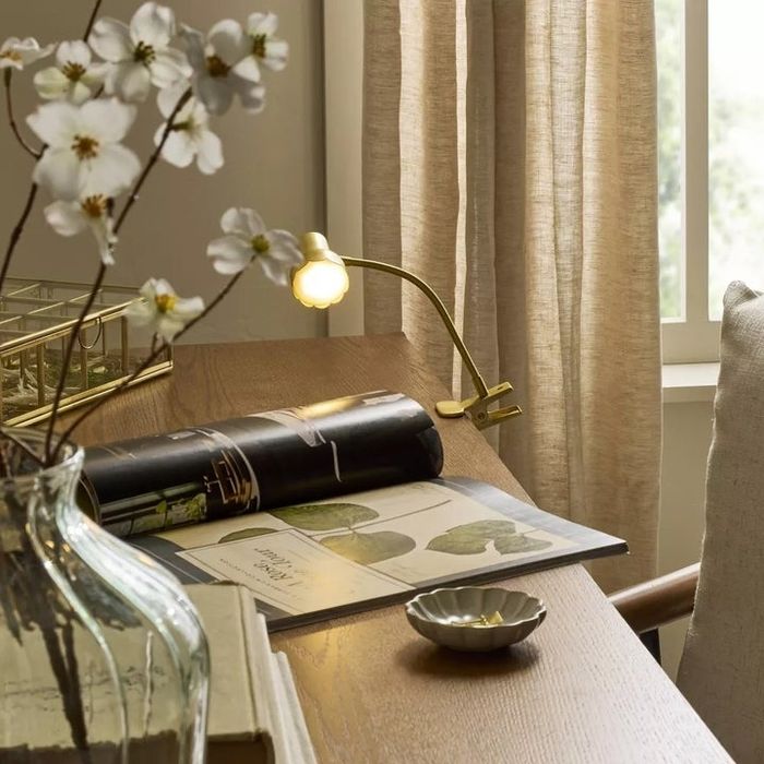 A modern wooden desk featuring an open magazine, a vase of white flowers, a small dish, and a reading lamp, all coming together to create a cozy shopping vibe.