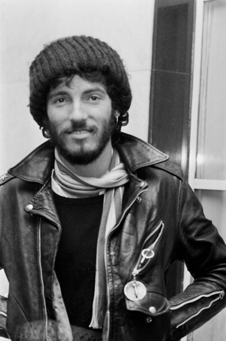 A person wearing a beanie and a leather jacket with a scarf, standing inside, offering a small smile to the camera