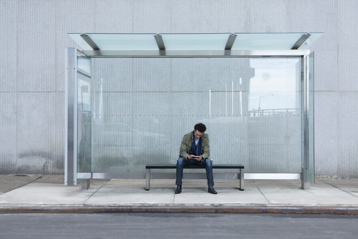 A person sitting alone on a bench at a glass bus stop, reading something in their hands