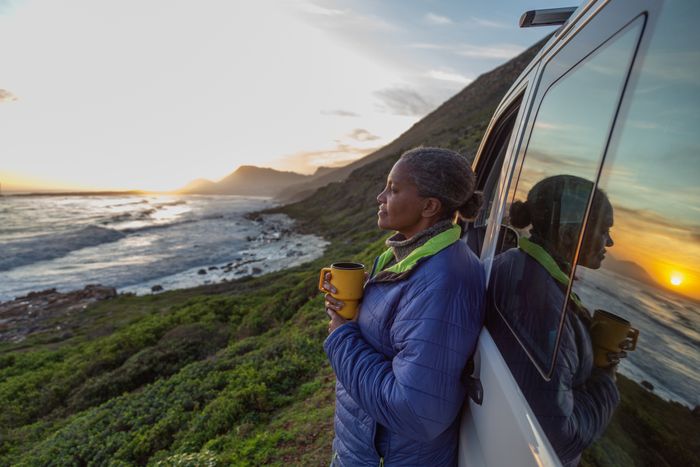 A person in a jacket leaning against a vehicle, holding a mug and watching an ocean sunset, with a rugged coastline in the background