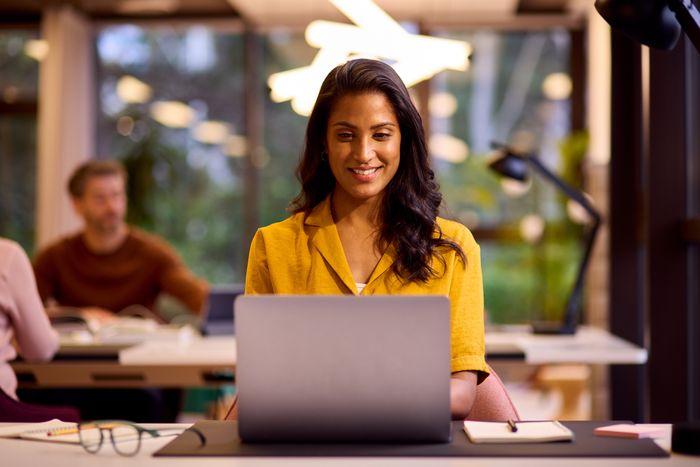 A person in a contemporary office setting, smiling and working on a laptop at a desk surrounded by notebooks and glasses.