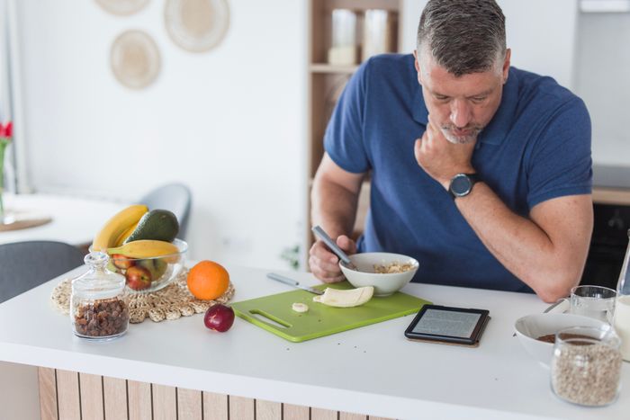 A man casually dressed, enjoying breakfast while reading a digital device at a kitchen counter surrounded by fruits and a cutting board.