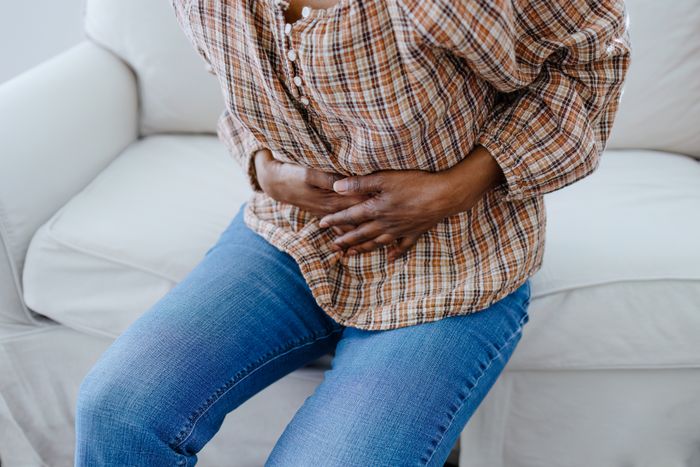 A person sitting on a couch, clutching their stomach in discomfort, wearing a plaid shirt and jeans.