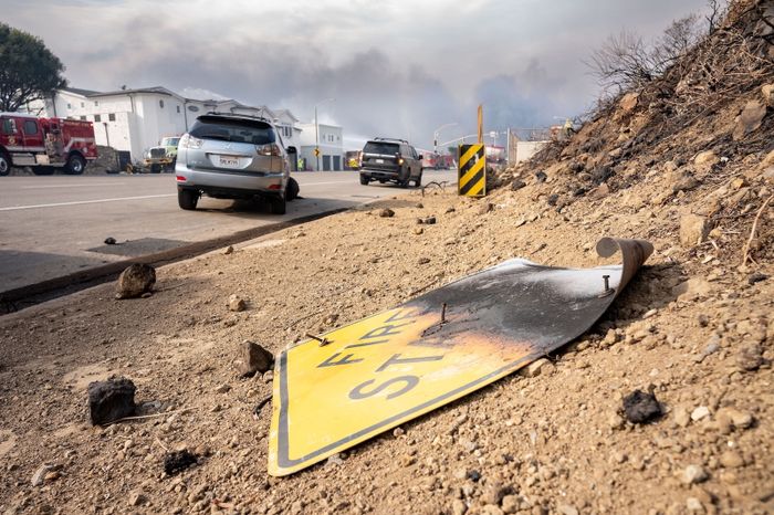 A fire station sign, now deformed, rests on a dirt slope with cars and emergency vehicles visible behind it