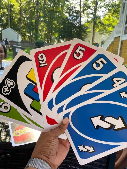A hand holding oversized UNO cards, including a Draw 4 Wild card, a 9, a 5, a 6, and a Reverse, all from different color sets. Trees and houses are seen in the background
