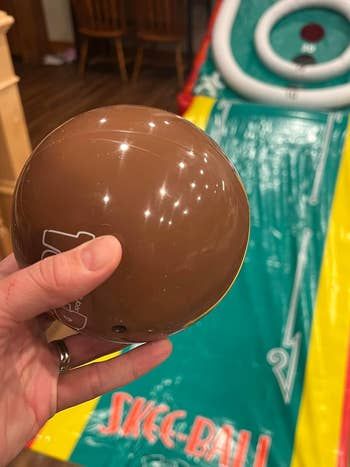 A hand holding a brown Skee-Ball with the Skee-Ball game setup in the background