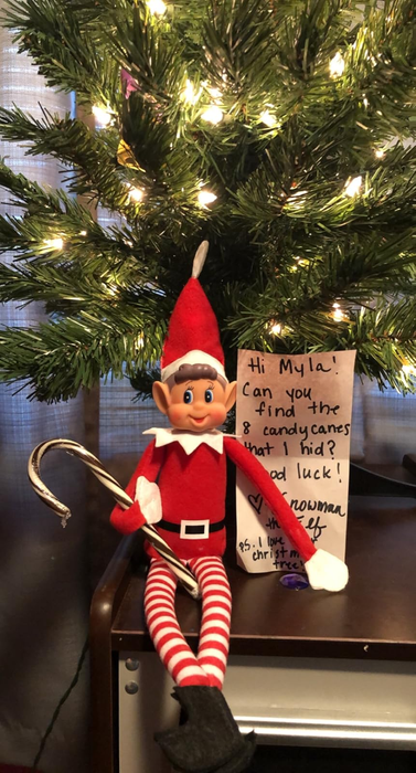 Elf doll clutching a candy cane, sitting next to a note beneath a Christmas tree
