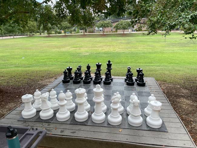 A life-sized outdoor chessboard with black and white pieces arranged for a game in a park