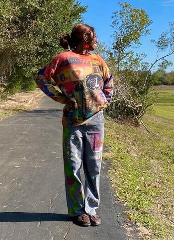 Person strolling on a paved path, wearing a vibrant sweater and patterned pants, surrounded by trees and a grassy field
