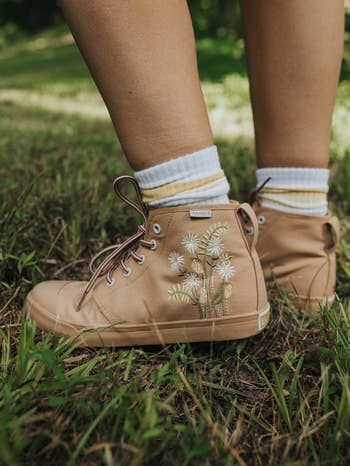 A close-up of legs wearing white and yellow striped socks, paired with light tan high-top sneakers adorned with white daisy embroidery, standing on grass.